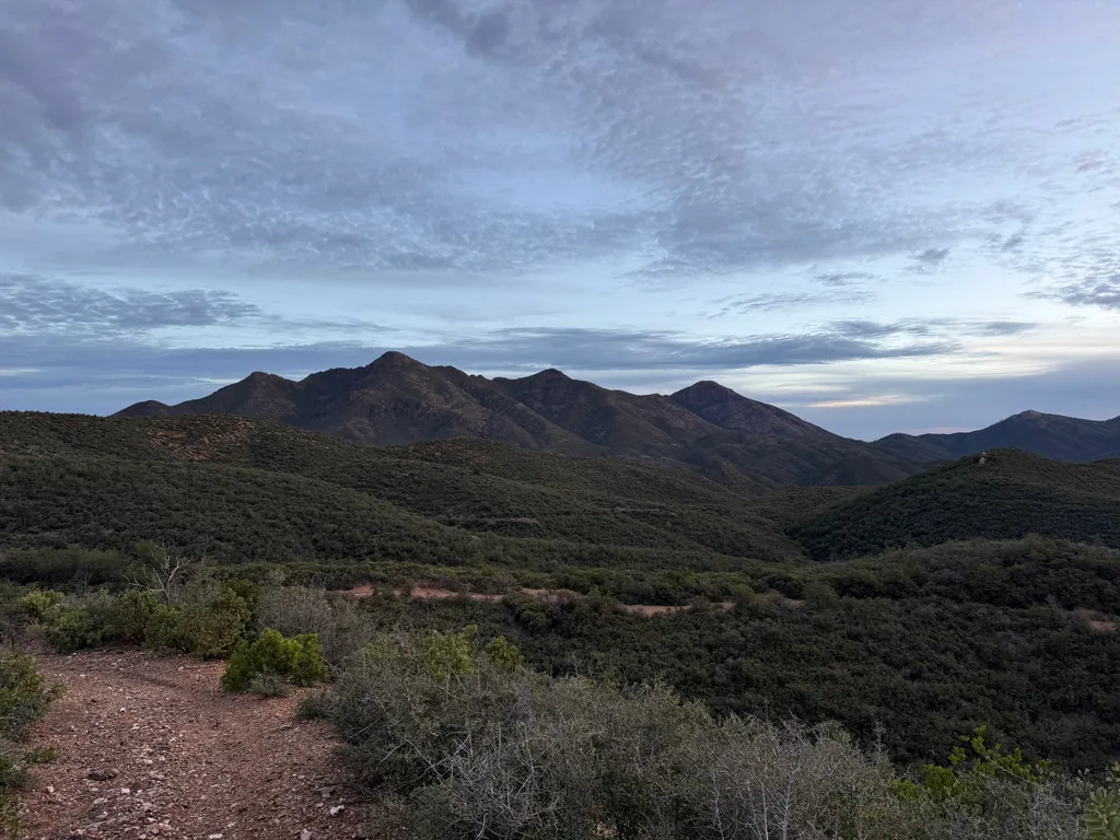 Serrated dark peak above rolling chaparral ridges, rippled grey cloud with a faint pale band at the horizon, gravel trail edge in the foreground