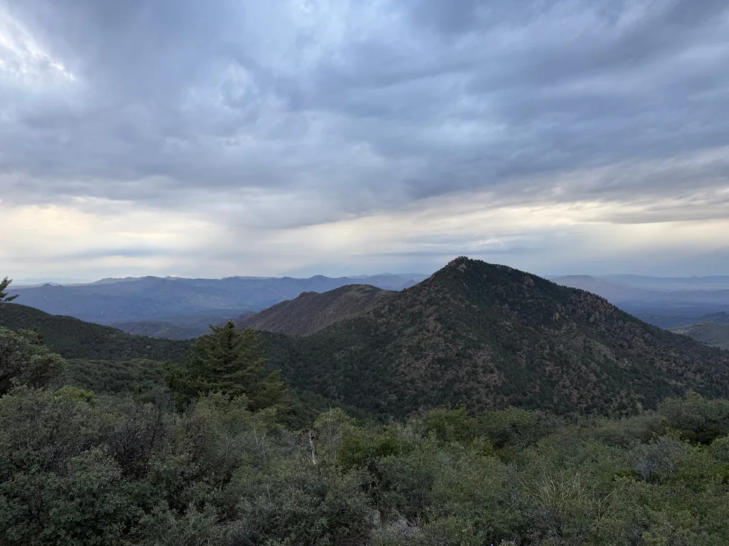 Cone-shaped wooded peak rising above darker ridges, under a thick grey cloud layer with pale light beneath; scrubby brush in the foreground