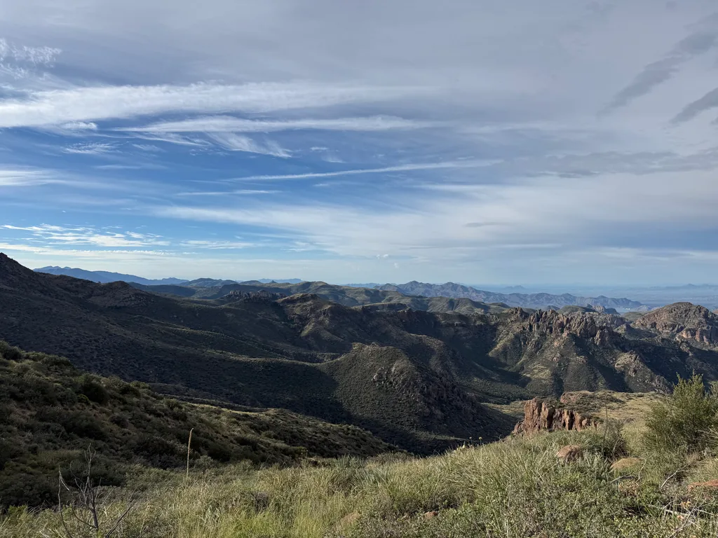 Green desert ridges stepping down to distant ranges, under streaked cirrus cloud; grass and shrubs in foreground