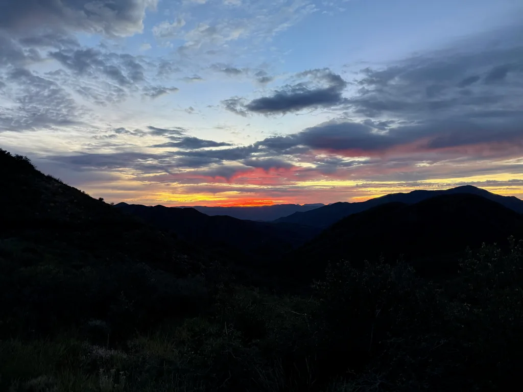 Narrow orange and red band burning between dark ridgelines, with grey cloud layers above and black brushy slopes below