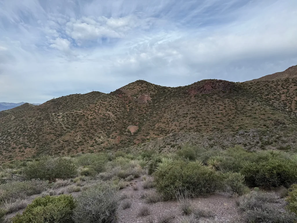 Brush-covered desert ridge with patches of red-orange exposed rock, gravel and yucca in the foreground under flat gray cloud