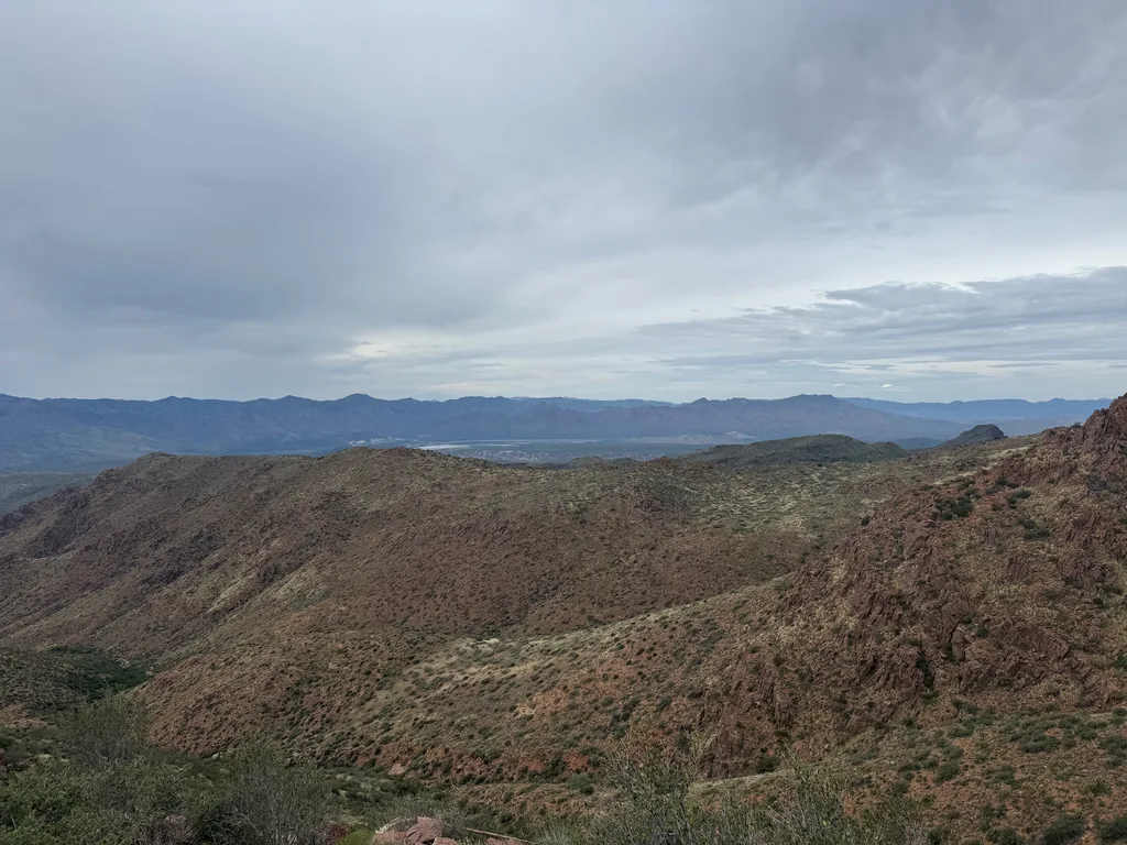 Rust-red rocky ridges dropping into a canyon, with a pale strip of water and hazy mountains beyond under solid grey cloud