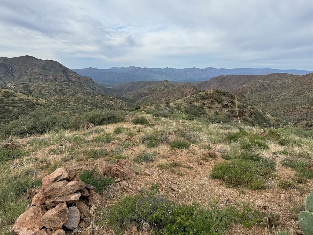 Stacked red rock cairn on a grassy shoulder, ridges fading into the distance under grey cloud