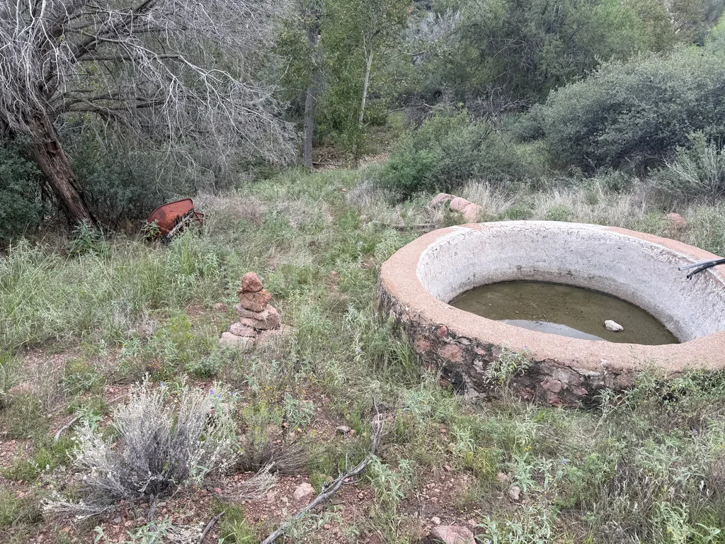 Circular stone-and-concrete tank holding green water, with a small stacked-rock cairn and rusted metal debris in dry grass nearby