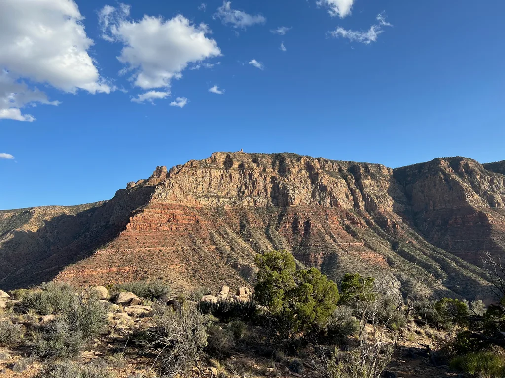 Tan cliff band over red and green terraced slopes, sunlit, with juniper and sagebrush in front