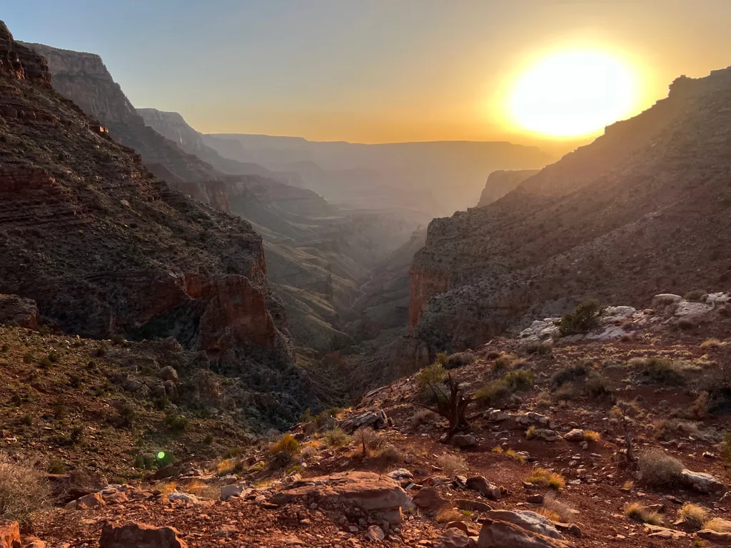 Sun low over a deep side canyon, red cliff bands in shadow, rocky slope with dry shrubs in front
