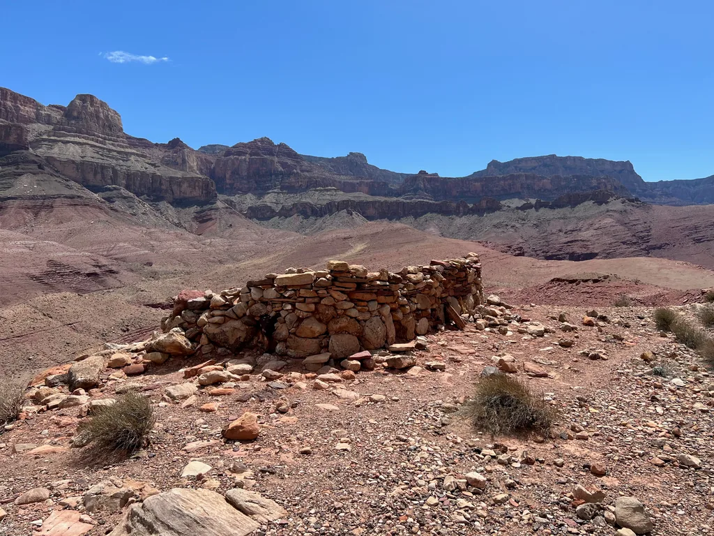 Waist-high dry-stacked stone wall enclosing a rectangle on a red gravel bench, layered canyon cliffs behind