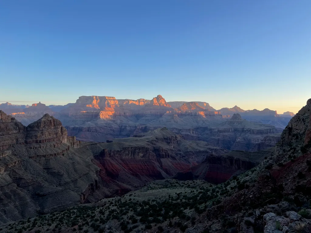 Distant flat-topped cliffs lit orange above canyon ridges still in blue shadow, clear sky overhead