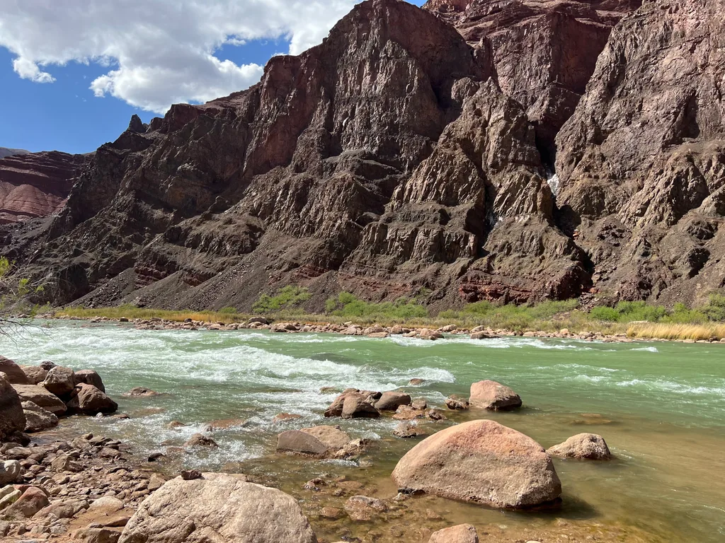 Pale green river running over rapids past rounded pink boulders, below dark red cliffs