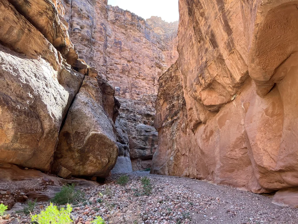A pale gray boulder wedged against red sandstone walls in a narrow canyon, cobble wash underfoot