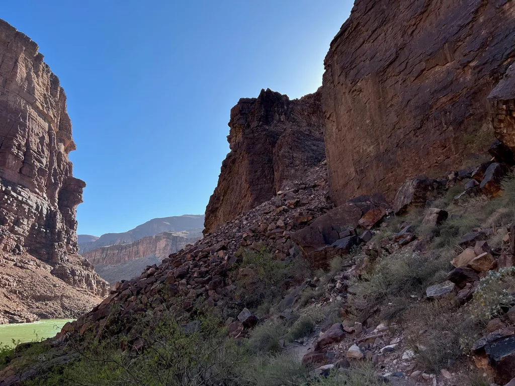 Steep boulder slope covered in scrub below a dark cliff, with green river water far below