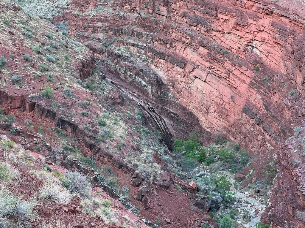 Steeply tilted grey rock layers meeting flat red cliff bands across a narrow side canyon, sparse shrubs on the slopes
