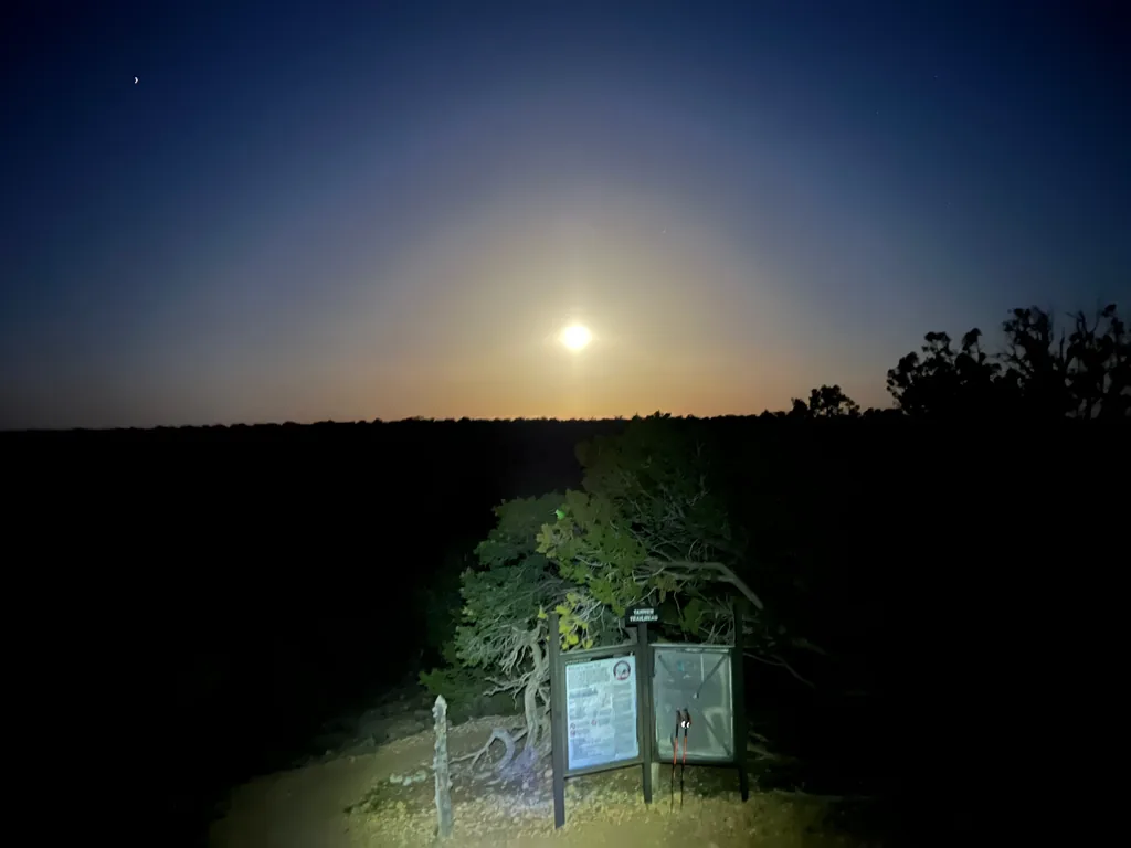 Bright moon low over dark treeline, with a lit information kiosk and small figure at a gravel trailhead below