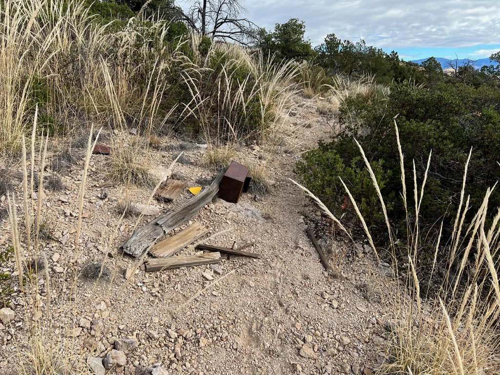 Splintered wooden post and rusted metal box lying beside a gravel trail through tall pale grass
