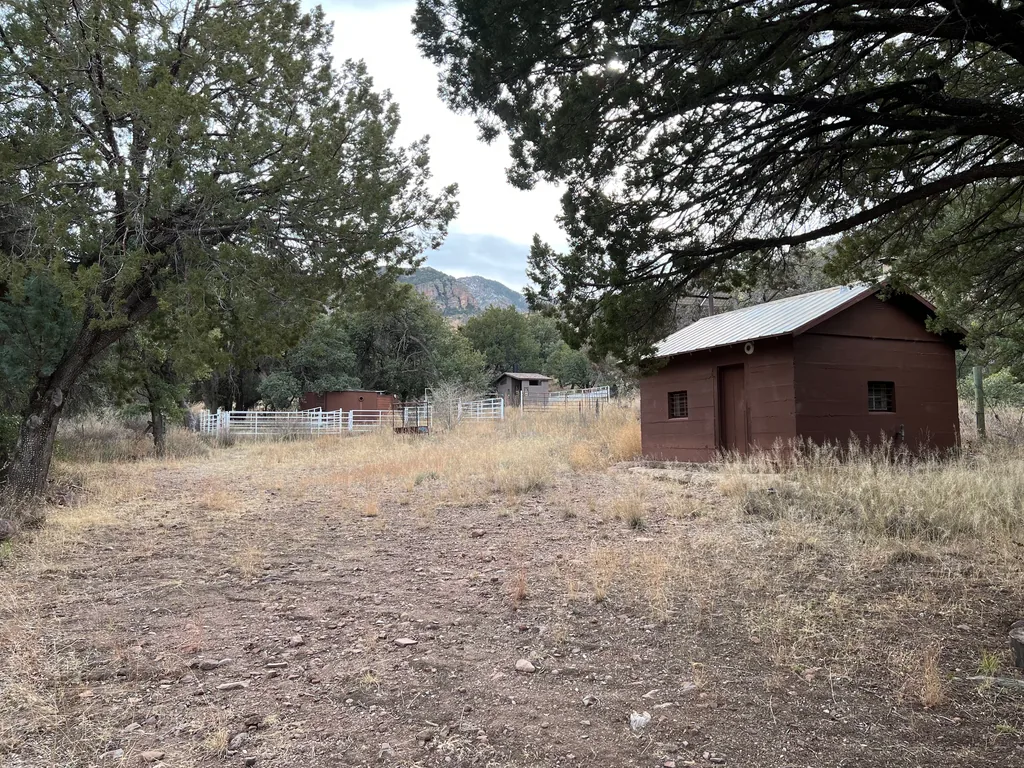 Small brown wood cabin with a metal roof beside dry grass, white pipe corral fence and a rocky peak behind