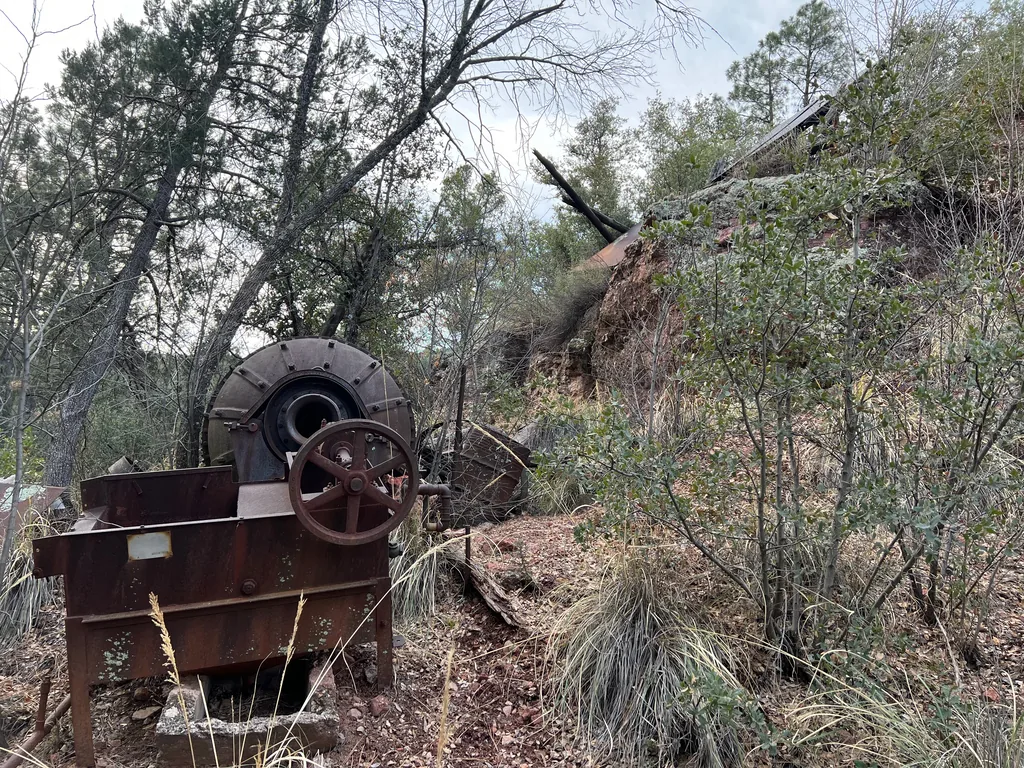 Rusted machine with a large flywheel and drum sitting in dry grass below a wooded slope
