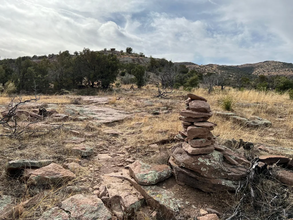 Waist-high cairn of flat pink lichen-spotted slabs beside a rocky trail through dry grass, low juniper hills behind