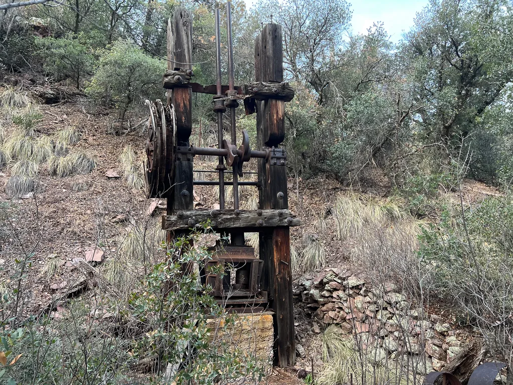 Weathered timber frame holding a rusted crankshaft and flywheel, brush and dry stone wall behind