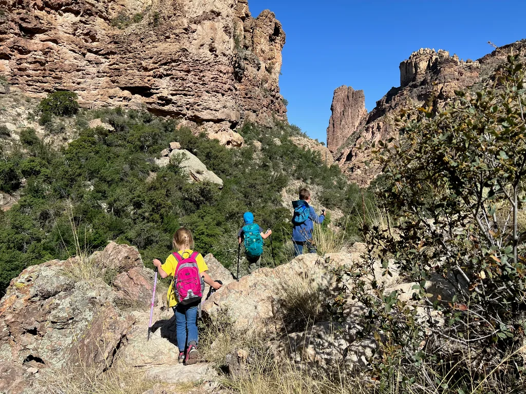 Three children with pink, teal and blue backpacks walk single file up a rocky trail below tall cliffs and a pointed rock spire