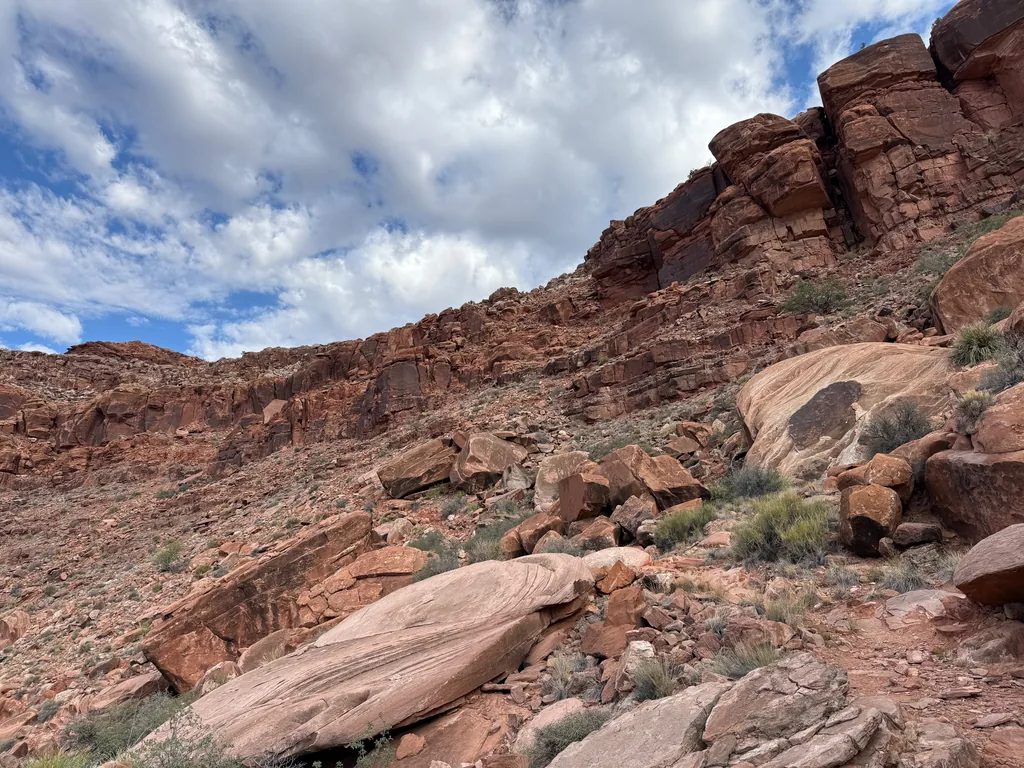 Smooth pink sandstone boulders with curved banding sit on a rubble slope below layered red cliffs, cumulus overhead