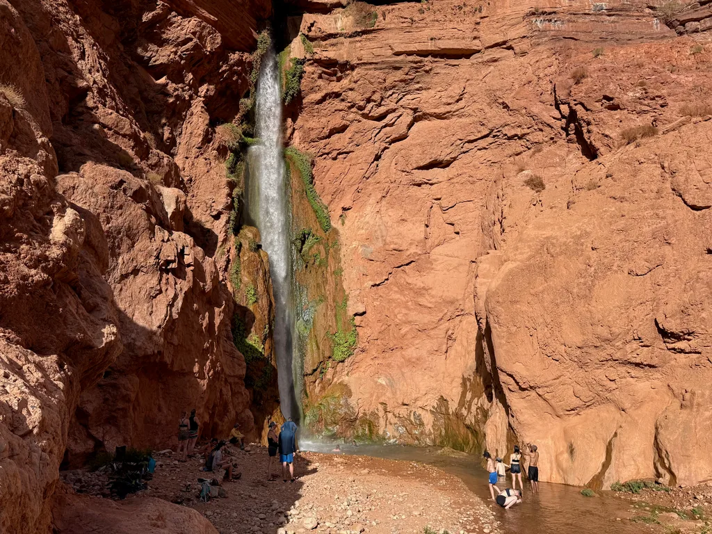 Thin waterfall drops down a red cliff streaked with green moss into a shallow pool where several people stand and wade
