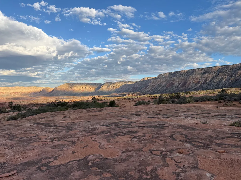 Flat red sandstone bench patterned with swirling dark lines, leading to a long banded cliff wall under scattered morning clouds