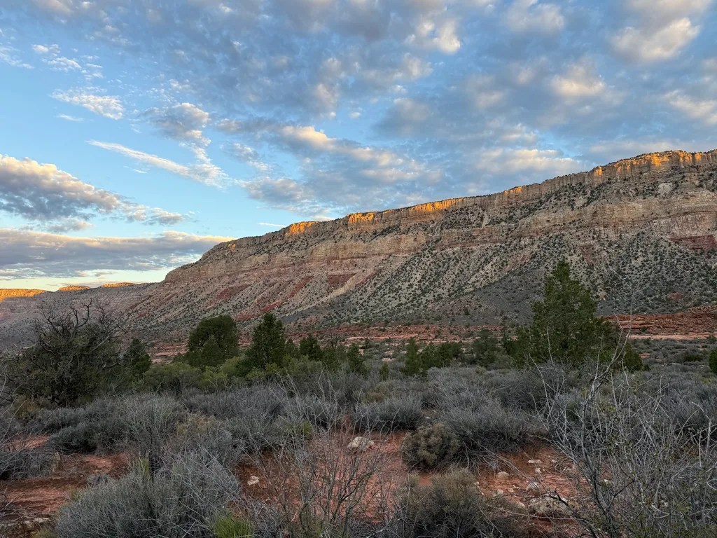 Orange sunlight strikes the top band of a long limestone cliff while slopes and sagebrush flats below stay in shade