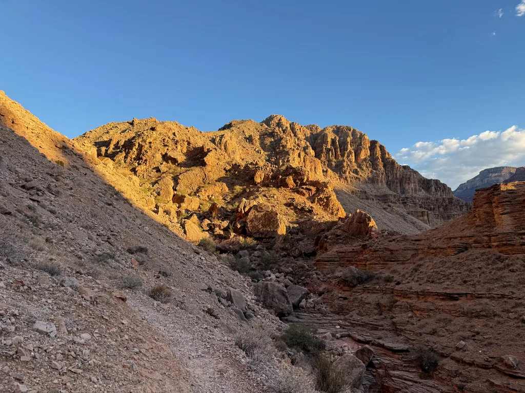 Sunlit orange cliffs above a drainage already in shadow, with layered red rock ledges at right under clear blue sky