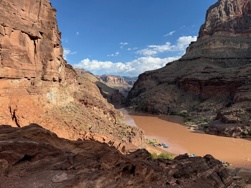 Cluster of small rafts moored on a sandbar along a wide brown river between red canyon walls