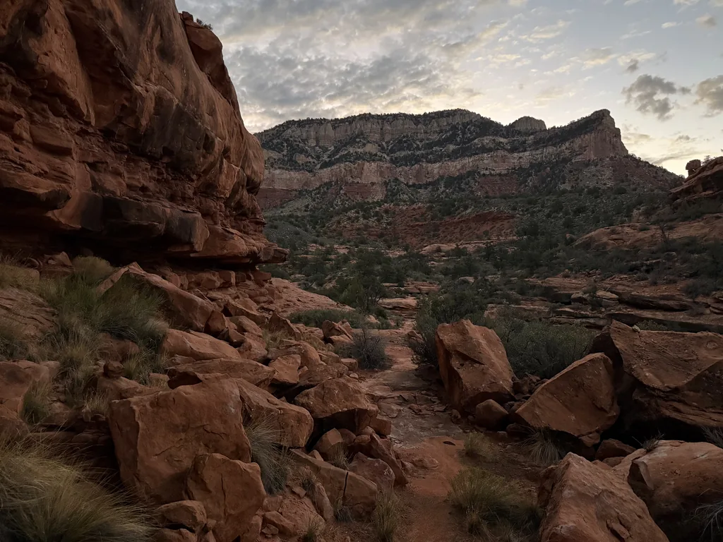 Trail threading between red sandstone boulders toward a banded cliff wall under grey dawn cloud