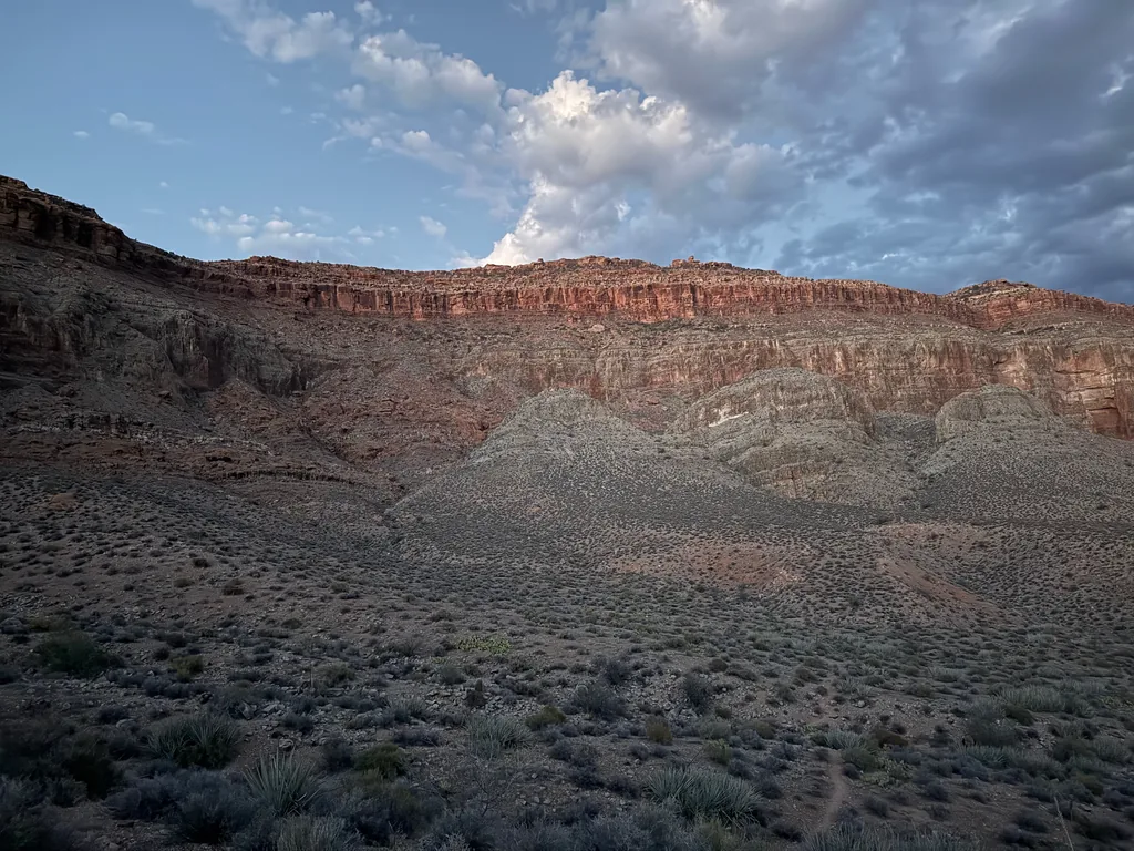 Red sandstone cliff band above grey mounds and sagebrush flats in dusk light, a tall white cloud behind