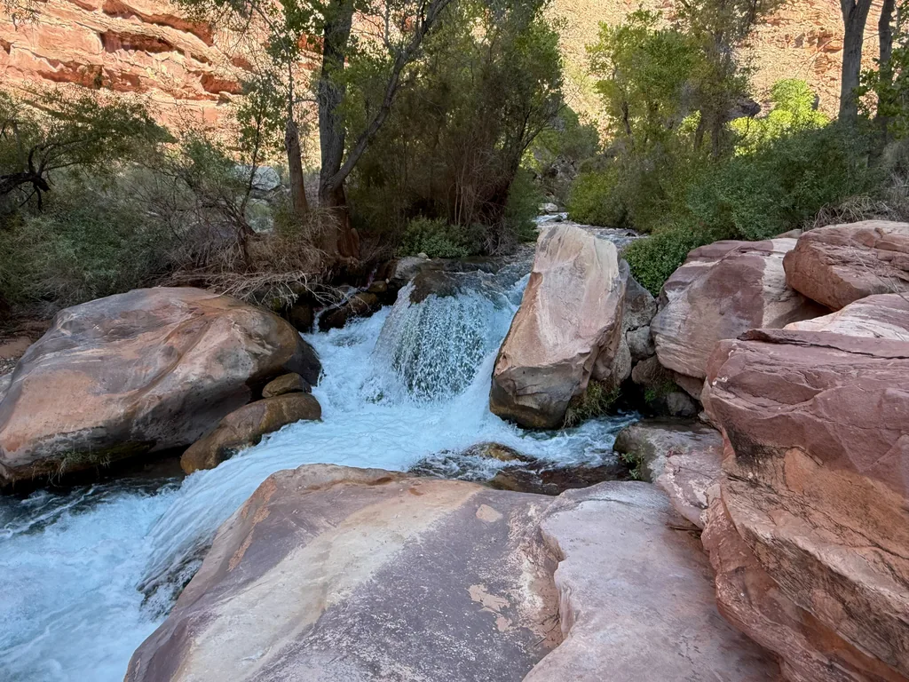 Whitewater pours through a narrow gap between rounded red sandstone boulders into a fast creek, green trees above