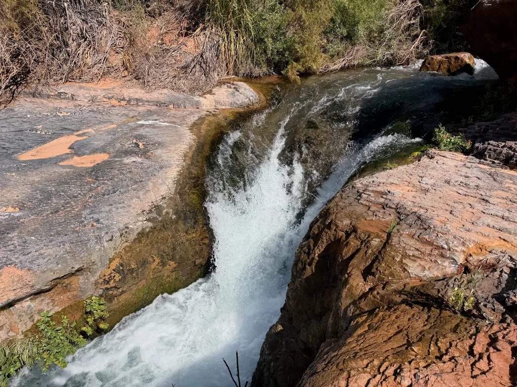 Creek water narrowing into a white chute through a gap between smooth red sandstone slabs, green weeds above