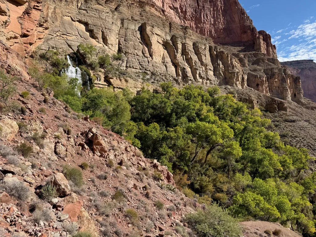 Thin white waterfall emerging partway down a tan limestone cliff, with bright green cottonwoods below and a red rocky slope in front