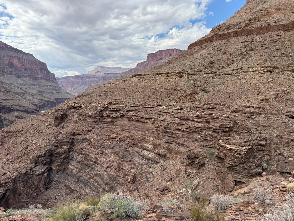 Thin red rock layers curving downward across a canyon wall, with a flat-topped butte and cloudy sky beyond