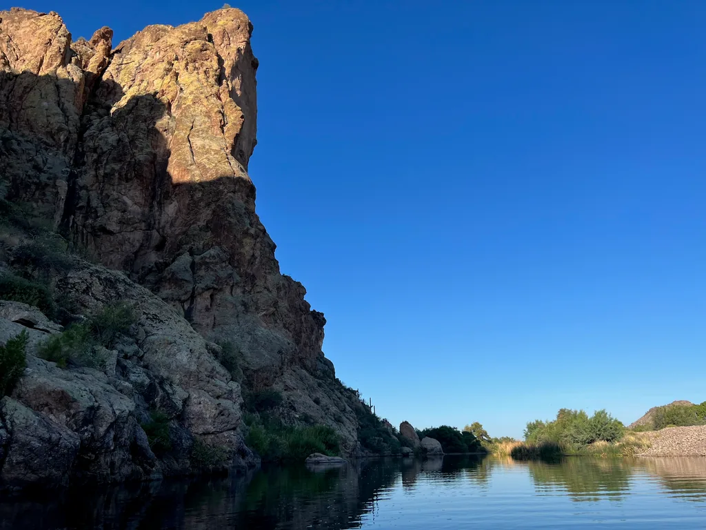 Tall rock cliff in shadow with a sunlit upper face, rising straight out of flat still water, a gravel bar and green trees on the far bank under a clear deep blue sky