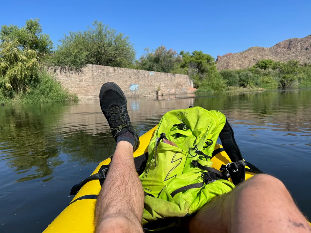 View past bare legs and one black water shoe propped on a yellow inflatable kayak, lime-green backpack between the knees, calm river ahead with a stone wall and rocky hills