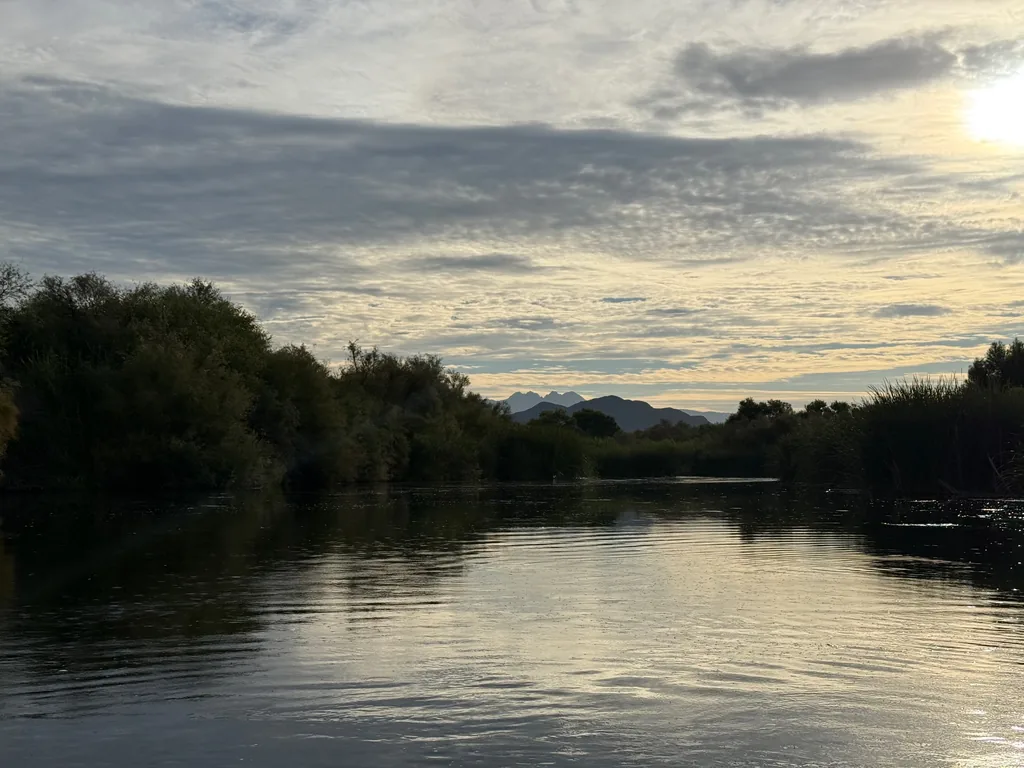 Flat river at water level, dark brush along both banks, the jagged crown of Four Peaks low on the horizon under a rippled morning sky