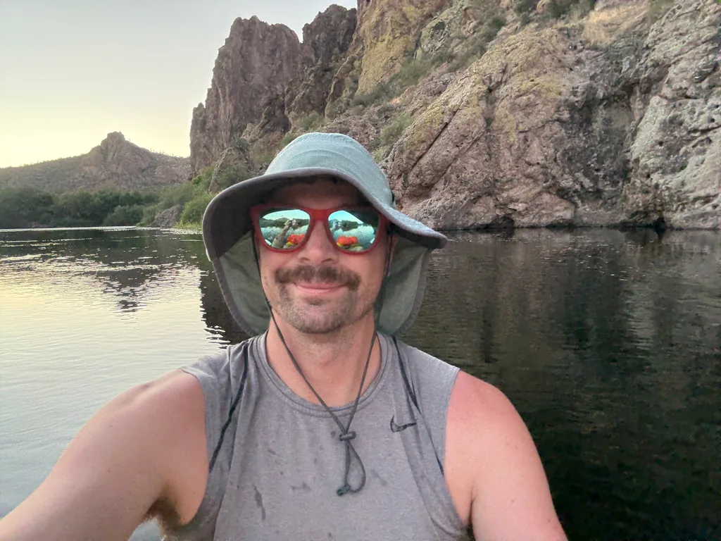Man in a wide-brimmed sun hat and red mirrored sunglasses smiling at the camera, cliffs and still water behind him