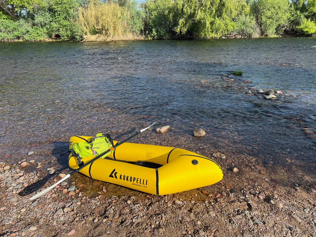 Yellow inflatable packraft beached on cobbles with a lime-green backpack inside and a black paddle laid across it, shallow river and willows behind